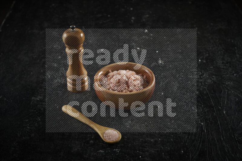 A wooden bowl and spoon filled with coarse pink himalayan salt and a wooden grinder beside them on black background