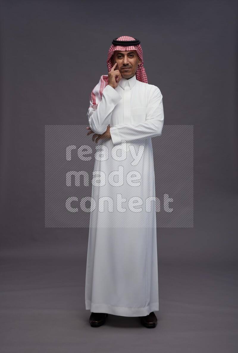 Saudi man wearing thob and shomag standing interacting with the camera on gray background