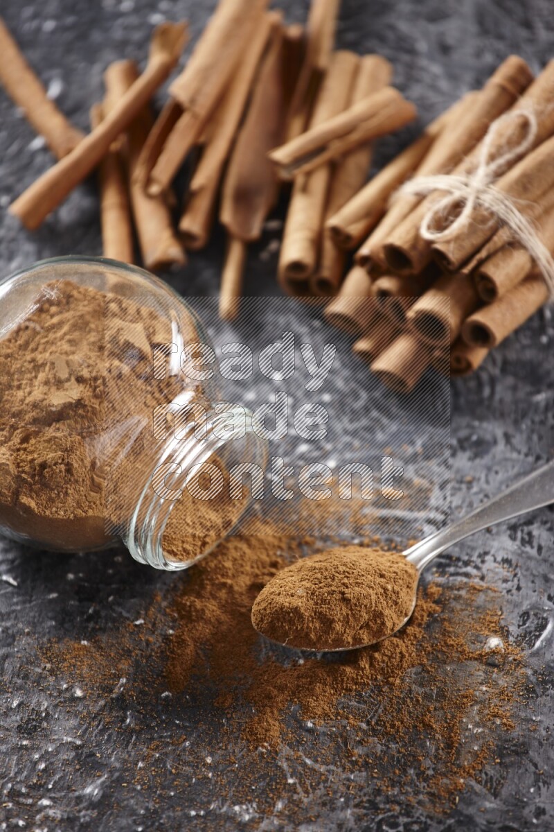 Herbal glass jar full cinnamon powder flipped and a metal spoon full of powder, cinnamon sticks stacked and bounded in the back on textured black background in different angles