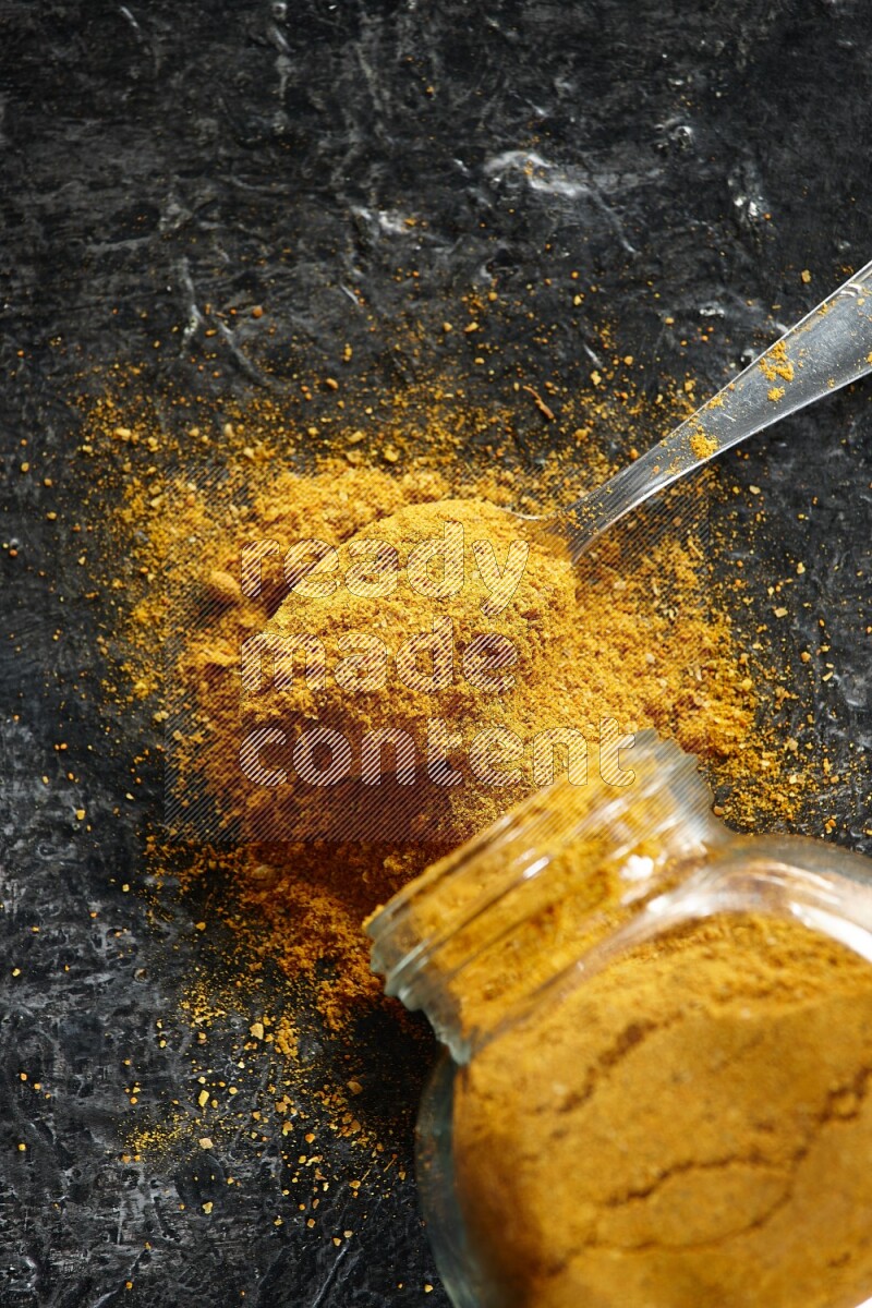 A flipped glass spice jar and a metal spoon full of turmeric powder and powder spilled out of it on textured black flooring