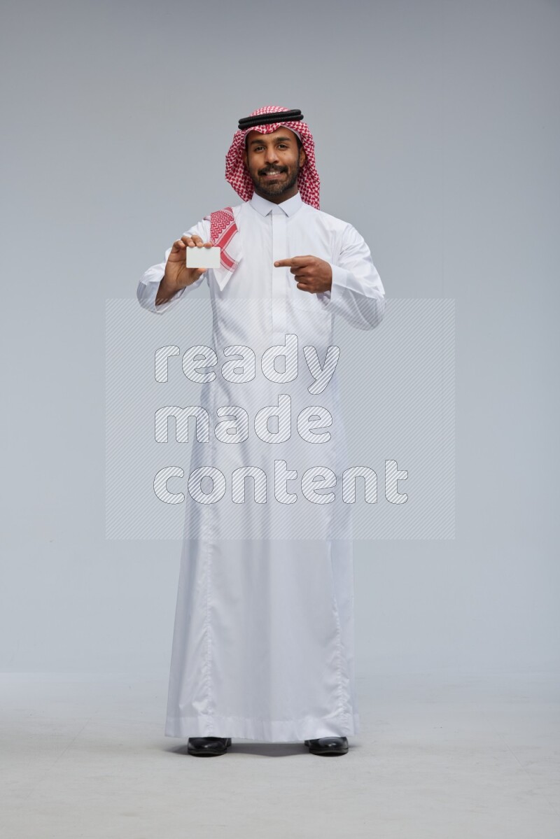 Saudi man wearing thob and shomag standing holding ATM card on gray background