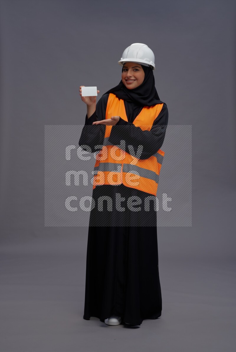 Saudi woman wearing Abaya with engineer vest standing holding ATM card on gray background