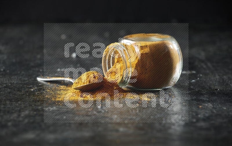 A flipped glass spice jar and a metal spoon full of turmeric powder and powder spilled out of it on textured black flooring