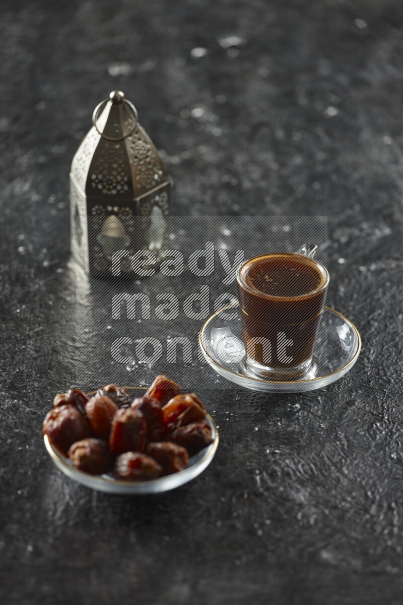 A silver lantern with different drinks, dates, nuts, prayer beads and quran on textured black background