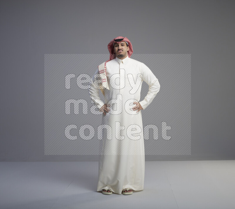 A Saudi man standing wearing thob and red shomag interacting with the camera on gray background