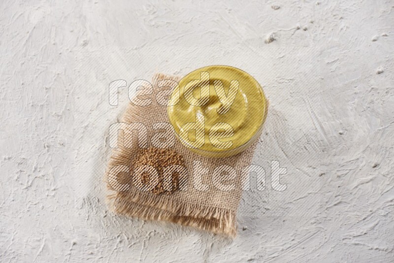 A glass bowl full of mustard paste set on a burlap piece with some mustard seeds spread on a textured white flooring