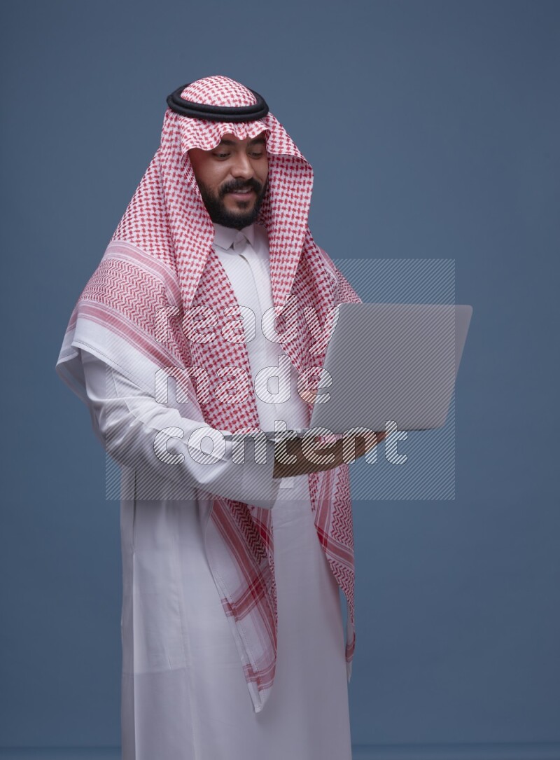 A man standing  with a laptop on Blue Background wearing Saudi Thob and Shomag