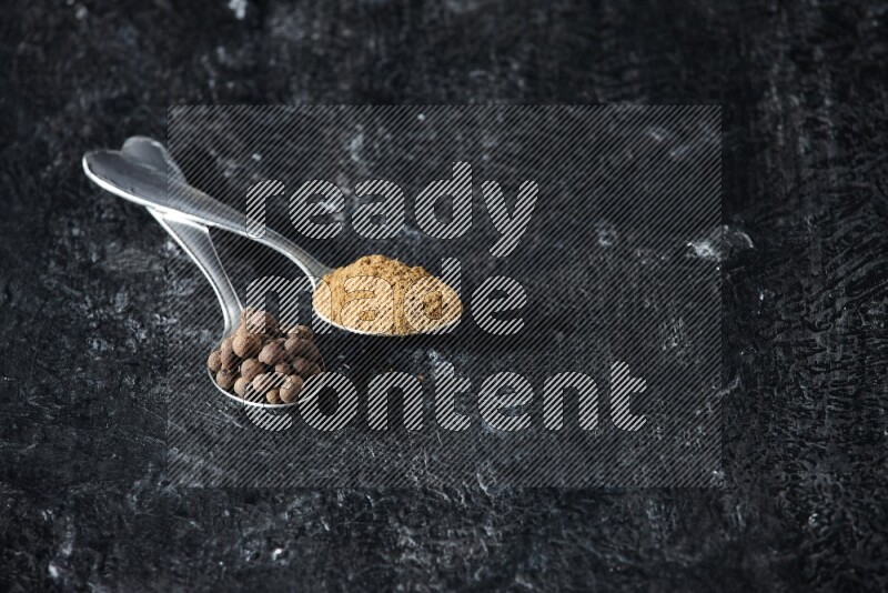 2 metal spoons full of allspice powder and whole balls on a textured black flooring