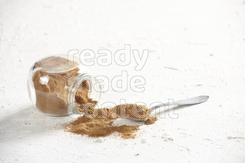 Flipped herbs glass jar full of cinnamon powder with a metal spoon full of powder on a textured white background
