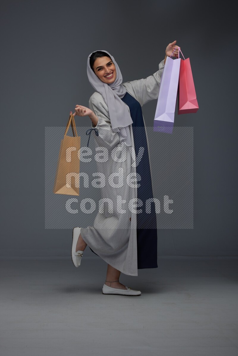 A Saudi woman wearing a light gray Abaya and head scarf standing and holding shopping bags on a grey background