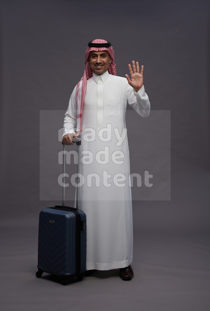 Saudi man wearing thob and shomag standing holding bag on gray background
