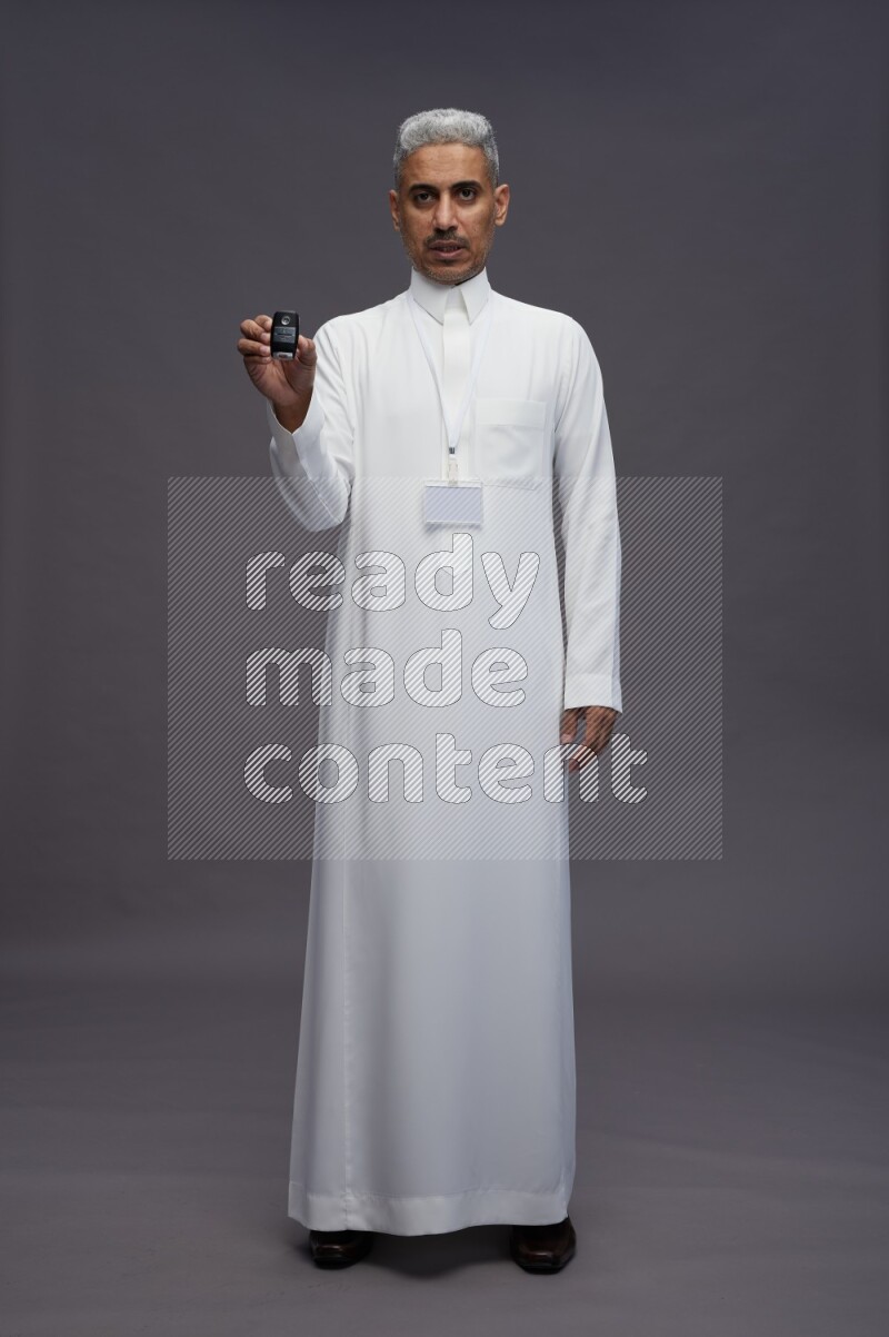 Saudi man wearing thob with neck strap employee badge standing holding key car on gray background