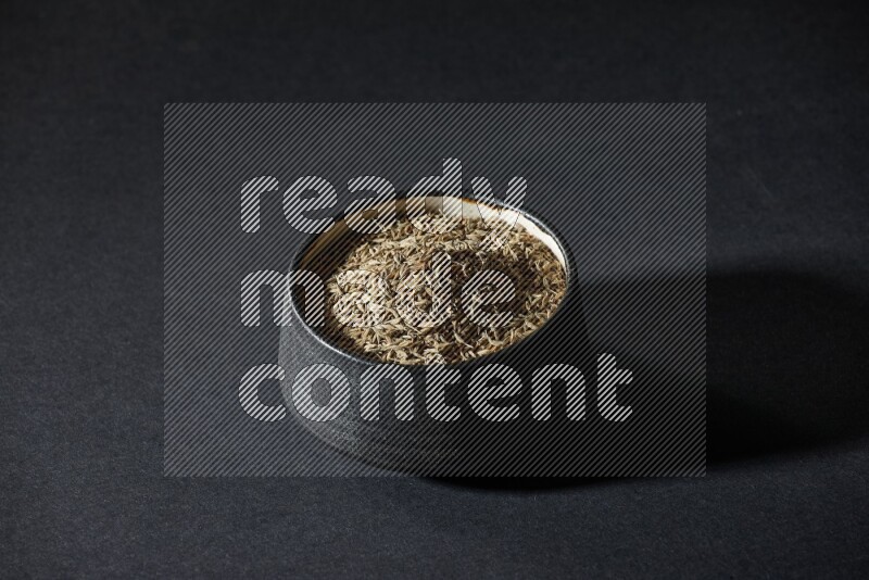 A black pottery bowl full of cumin seeds on a black flooring