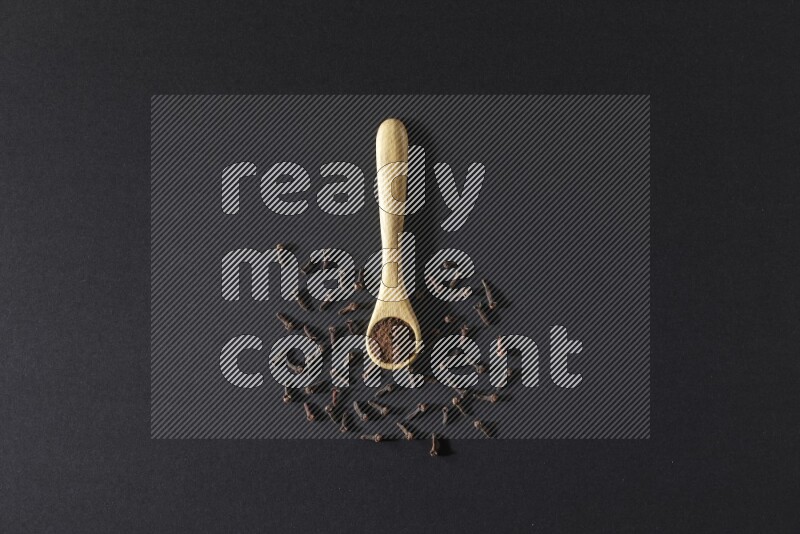 A wooden spoon full of cloves powder with spreaded whole cloves on a black flooring