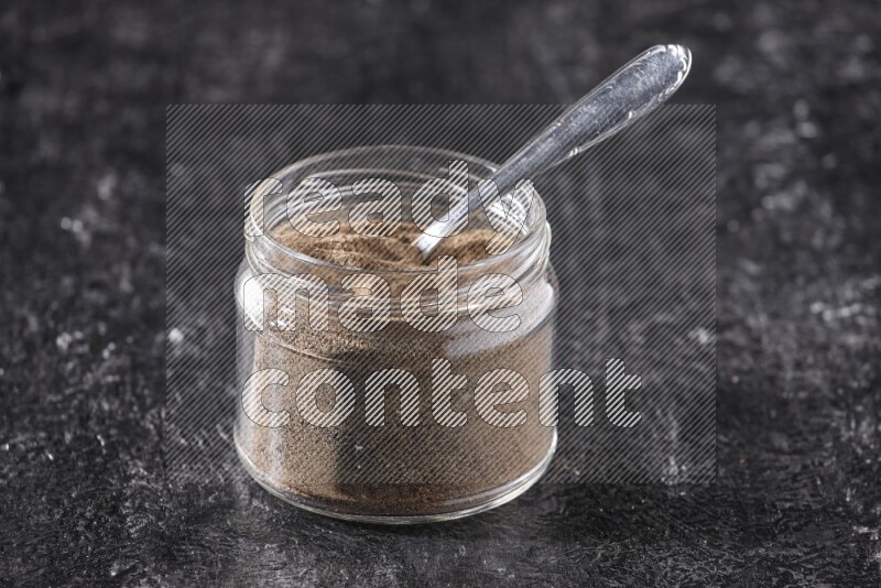 A glass jar full of black pepper powder and a metal spoon on a textured black flooring