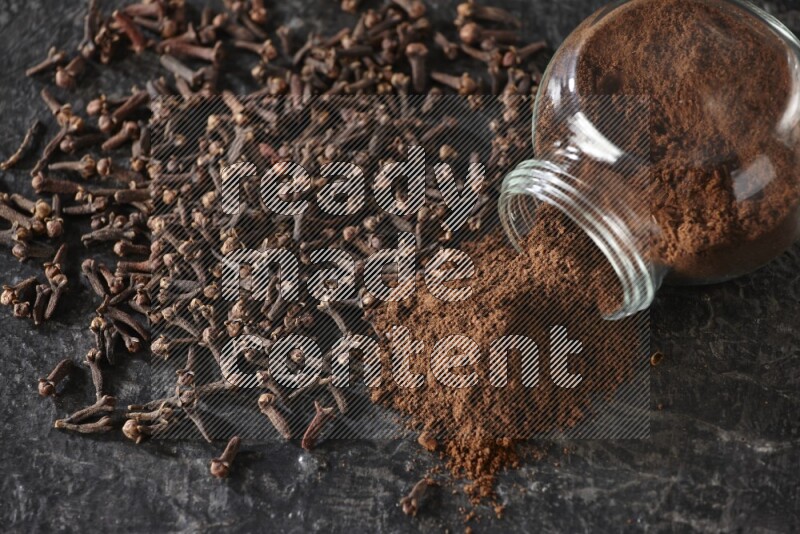 A flipped glass spice jar full of cloves powder and powder came out of it with cloves spread on textured black flooring