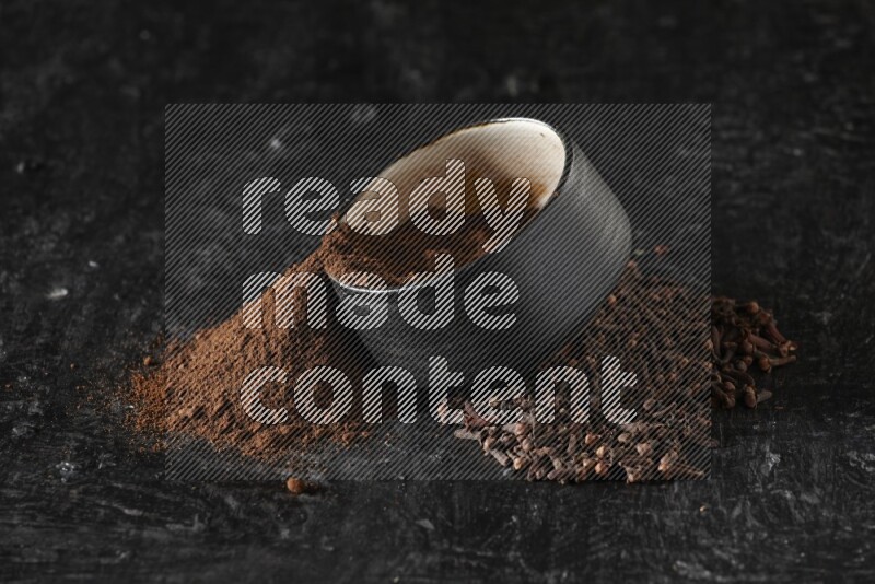 A black pottery bowl full of cloves powder spilled out of it with whole grains on a textured black flooring