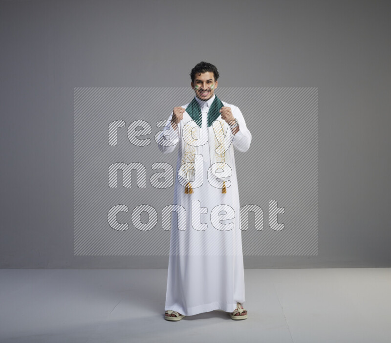 A Saudi man standing wearing thob and Saudi flag scarf with face painting on gray background