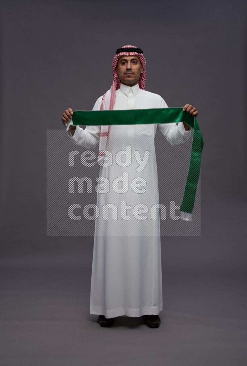Saudi man wearing thob and shomag standing holding Saudi flag on gray background