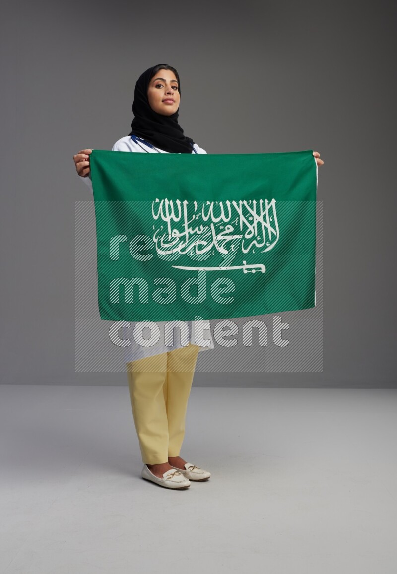 Saudi woman wearing lab coat with stethoscope standing holding Saudi flag on Gray background