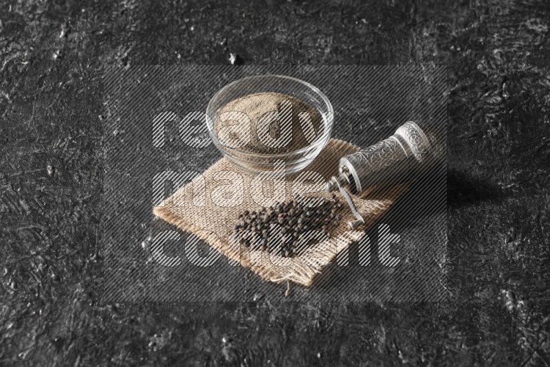 A glass bowl full of black pepper powder and black pepper beads on burlap fabric with a turkish metal pepper grinder on textured black flooring