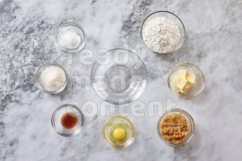 كوكيز خطوة بخطوة بمكوناتها، الدقيق، الزبدة، السكر البني، البيض، الفانيليا، السكر الأبيض، رقائق الشوكولاتة وصودا الخبز على خلفية رخام رمادي