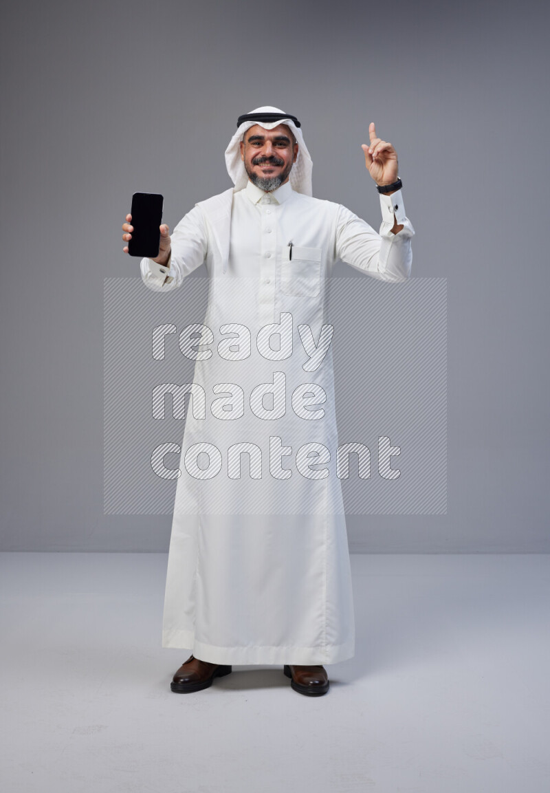 Saudi man Wearing Thob and white Shomag standing showing phone to camera on Gray background