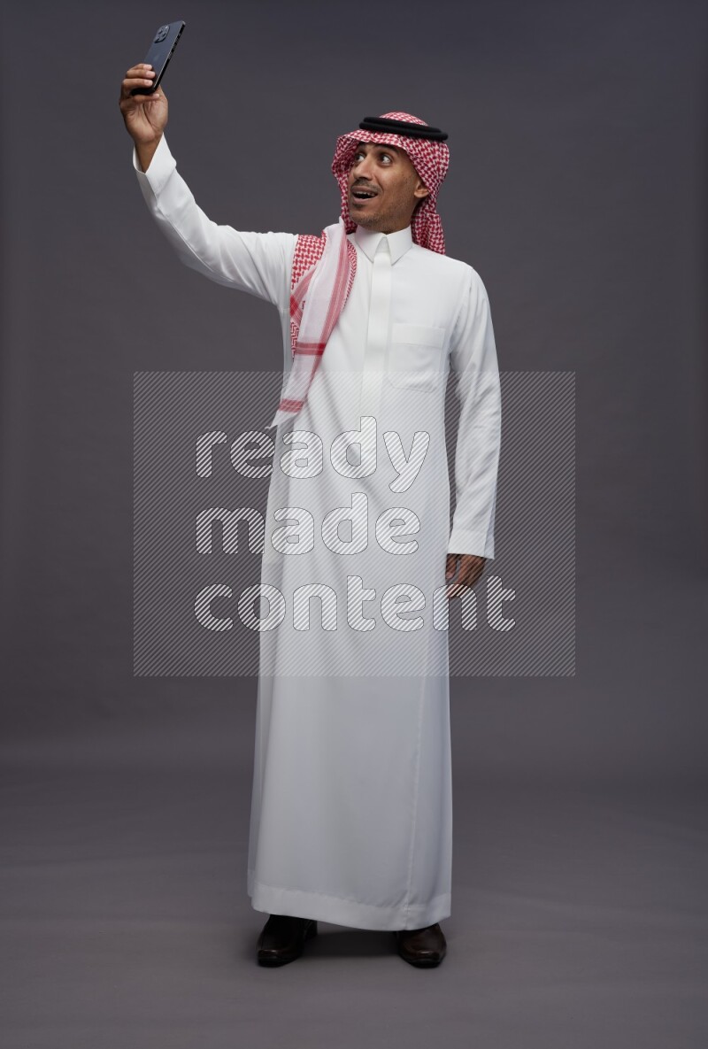 Saudi man wearing thob and shomag standing taking selfie on gray background