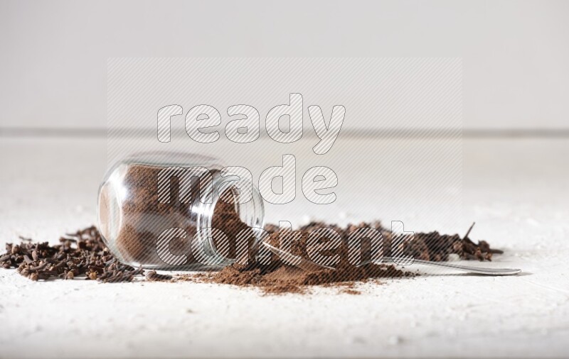 A flipped glass spice jar and a metal spoon full of cloves powder and powder came out of the jar with cloves spread on textured white flooring