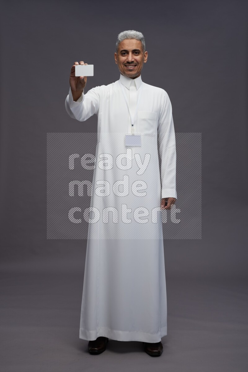 Saudi man wearing thob with neck strap employee badge standing holding ATM card on gray background