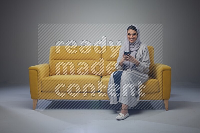 A Saudi woman wearing a light gray Abaya and white head scarf sitting on a yellow sofa and using her phone eye level on a grey background