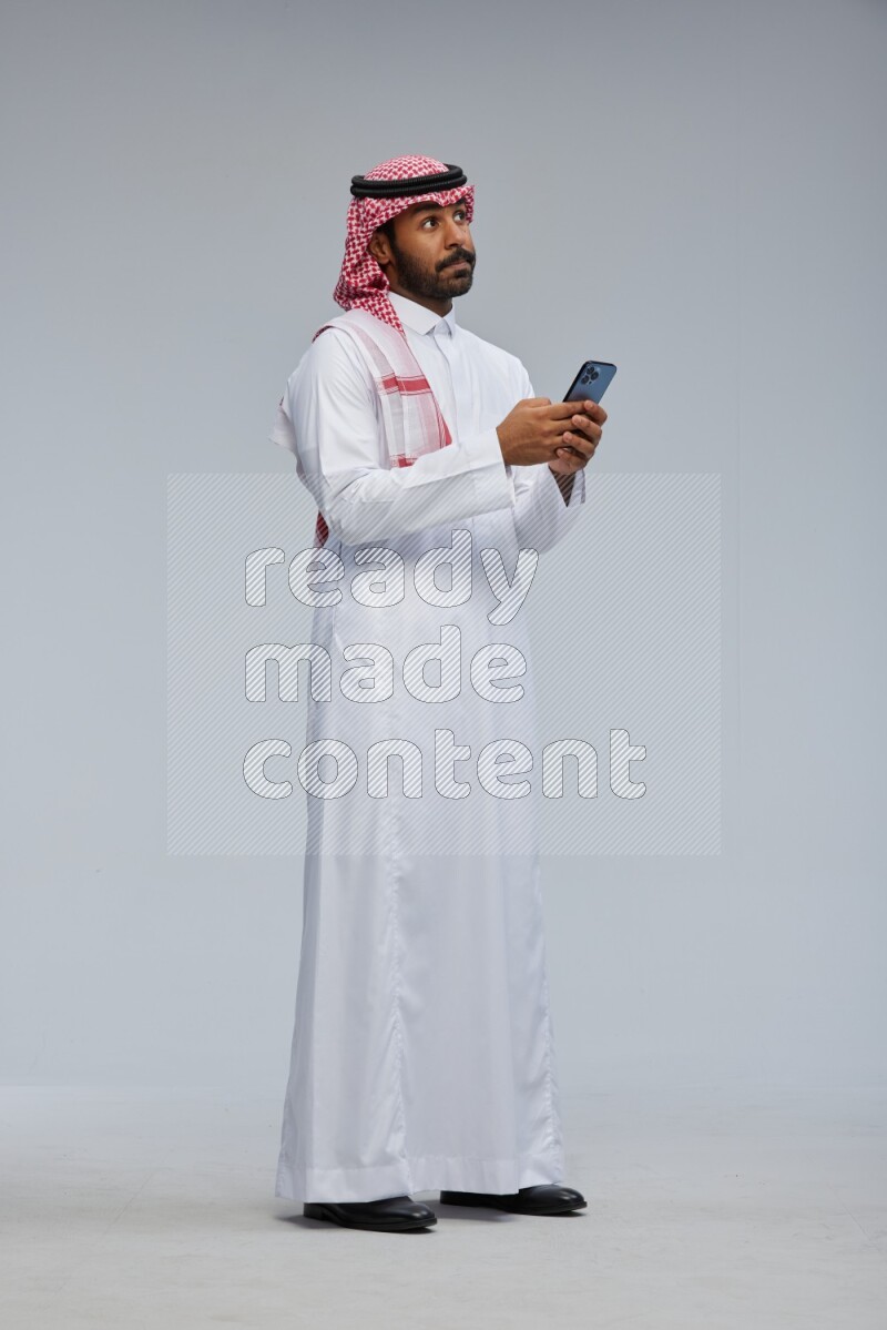 Saudi man Wearing Thob and shomag standing texting on phone on Gray background
