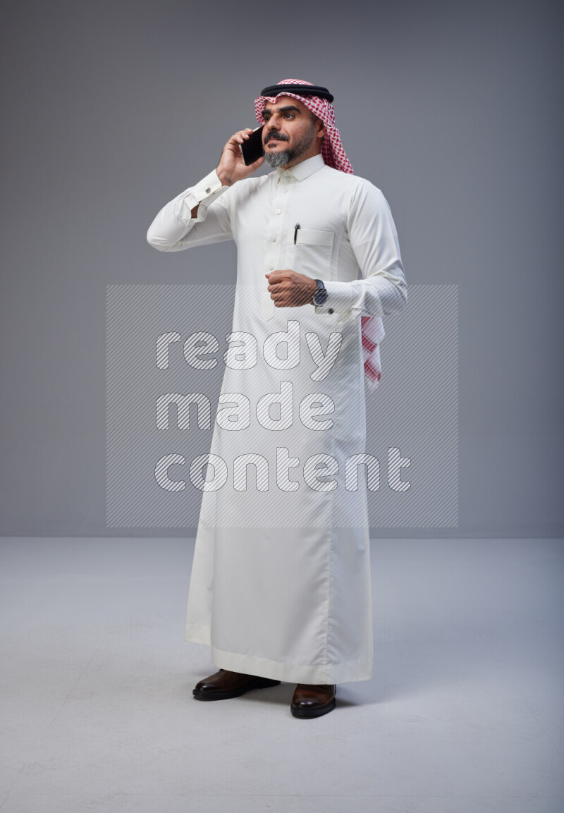 Saudi man Wearing Thob and red Shomag standing talking on phone on Gray background