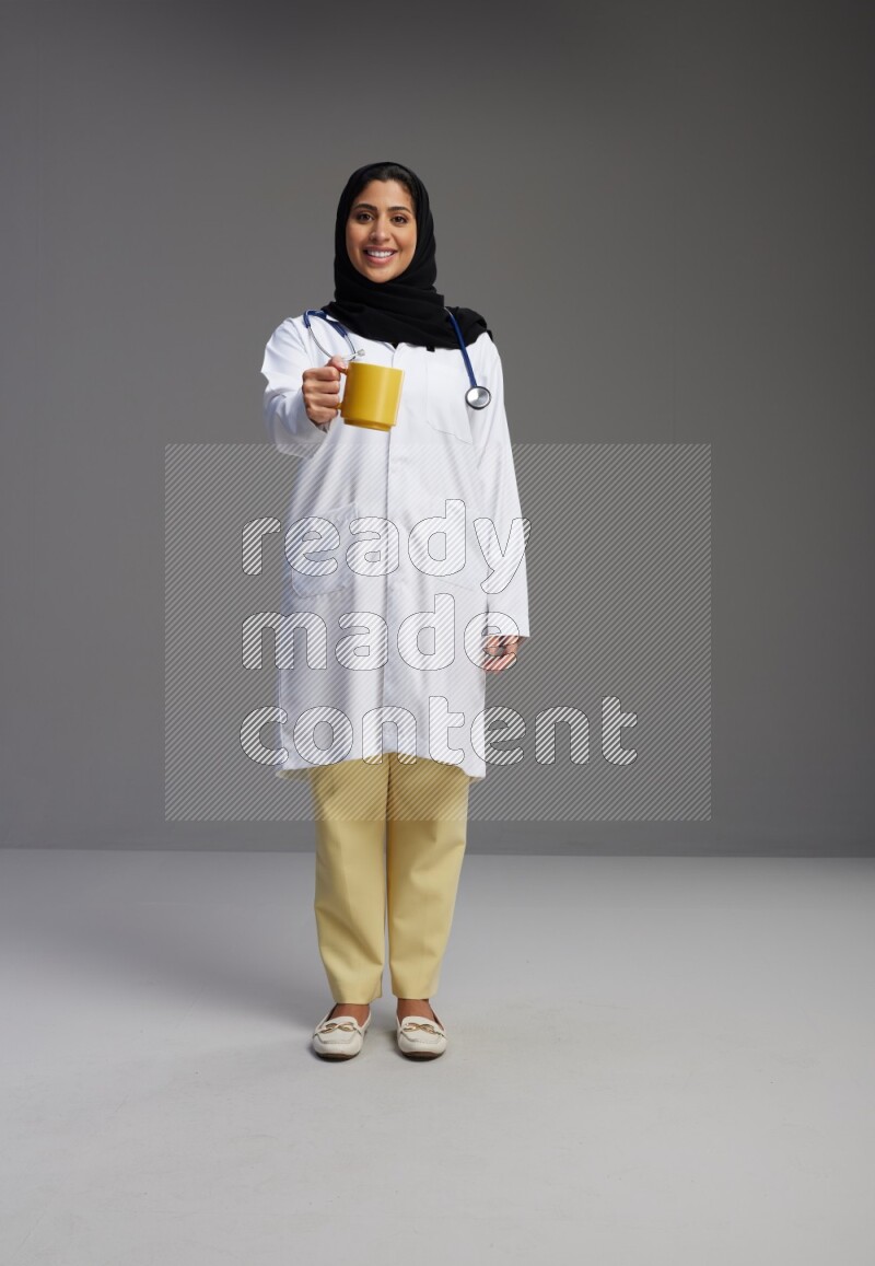 Saudi woman wearing lab coat with stethoscope standing holding mug on Gray background