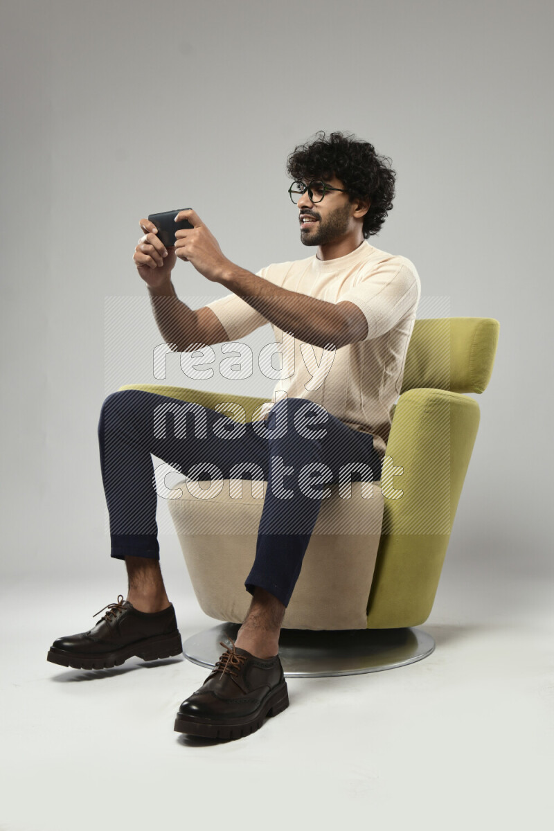 A man wearing casual sitting on a chair gaming on the phone on white background