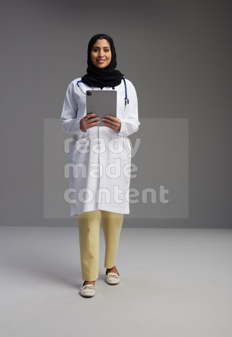 Saudi woman wearing lab coat with stethoscope standing working on tablet on Gray background
