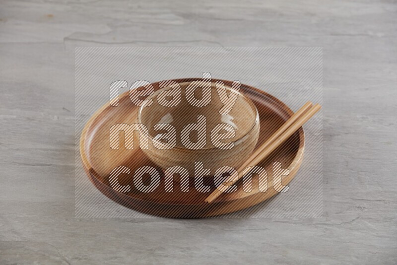 multi color pottery round bowl on top of brown wood round plate and wood chopsticks, on grey textured countertop
