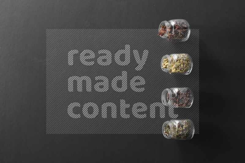 4 glass spice jars full of cloves, cardamom, basil and star anise on black flooring