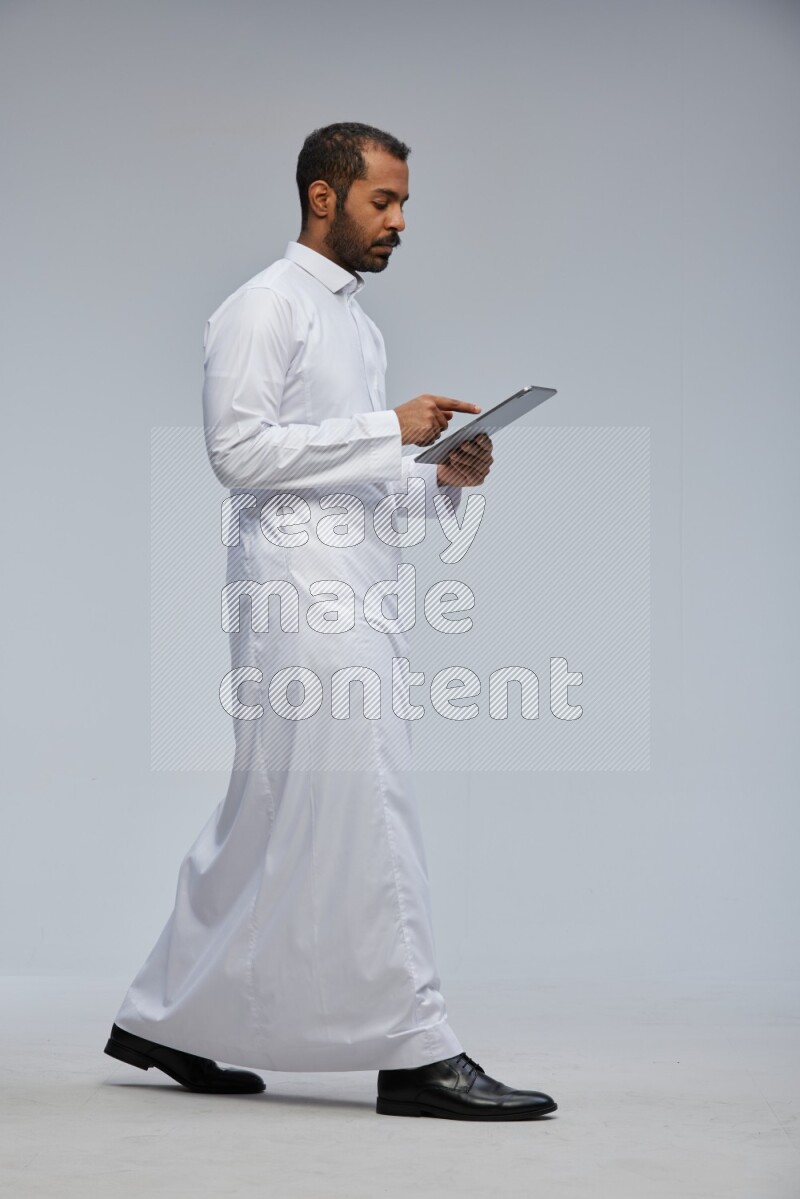 Saudi man wearing Thob standing working on tablet on Gray background
