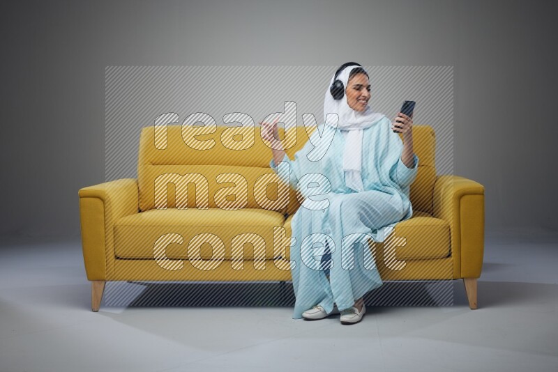 A Saudi woman wearing a light blue Abaya and a white head scarf sitting on a yellow sofa and holding an iPad while wearing headphone eye level on a grey background