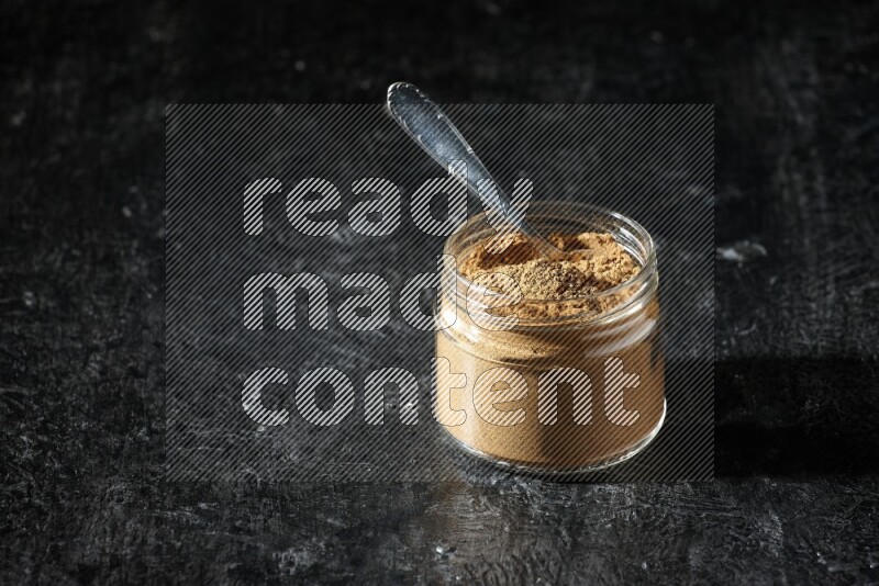 A glass jar and a metal spoon full of allspice powder on a textured black flooring