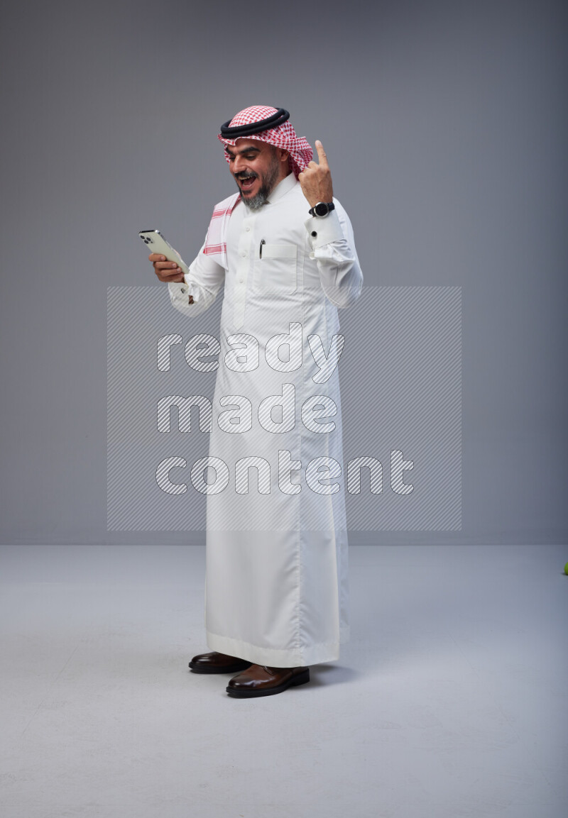 Saudi man Wearing Thob and red Shomag standing texting on phone on Gray background