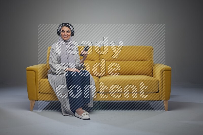 A Saudi woman wearing a light gray Abaya and head scarf sitting on a yellow sofa and using her phone while wearing headphones eye level on a grey background