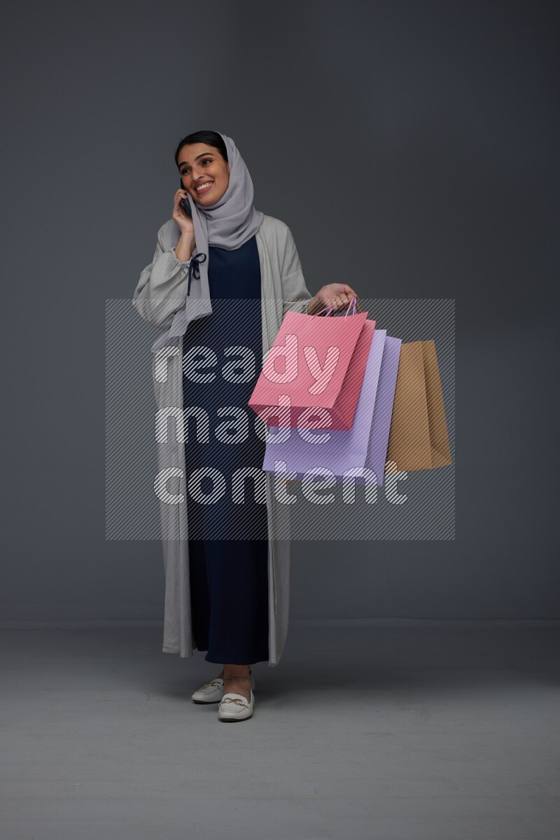 A Saudi woman wearing a light gray Abaya and head scarf standing and holding shopping bags making different poses eye level on a grey background
