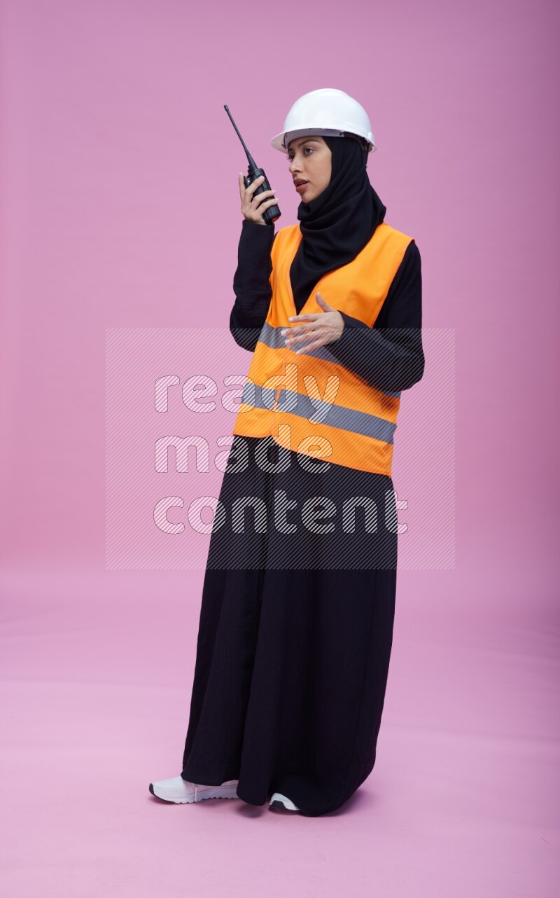 Saudi woman wearing Abaya with engineer vest and helmet standing holding walkie-talkie on pink background