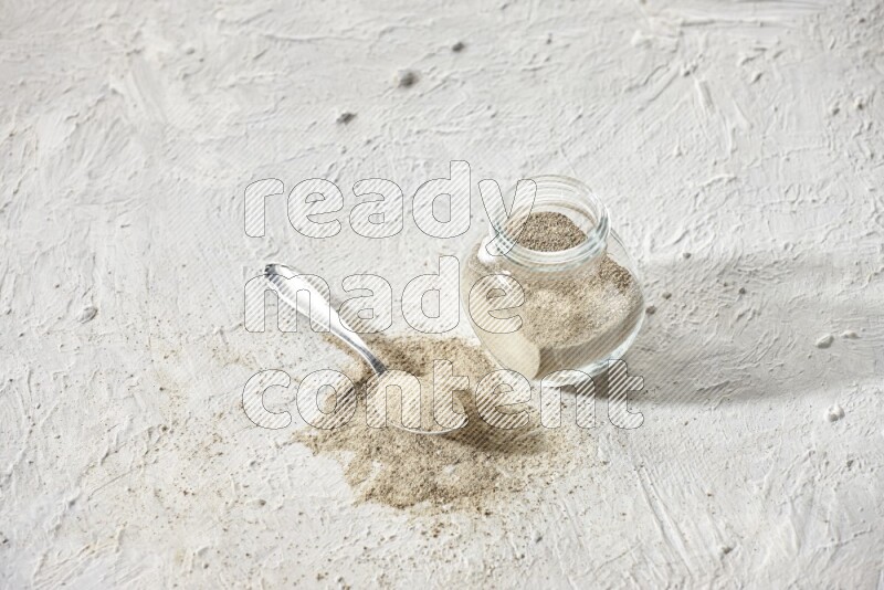 Herbal Glass jar full of white pepper powder and metal spoon full of powder on textured white flooring