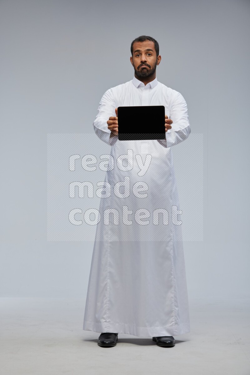 Saudi man wearing Thob standing showing tablet to camera on Gray background