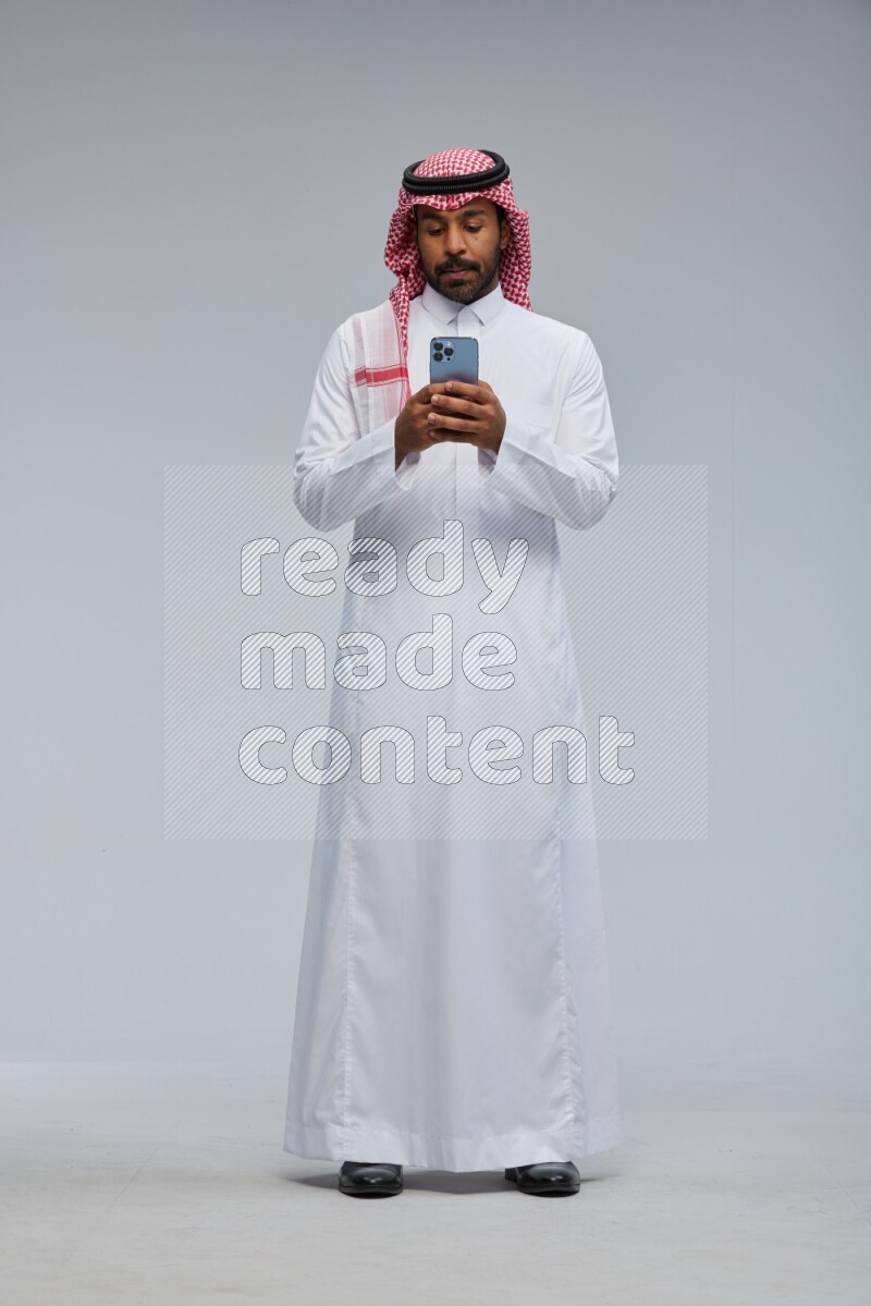 Saudi man Wearing Thob and shomag standing texting on phone on Gray background
