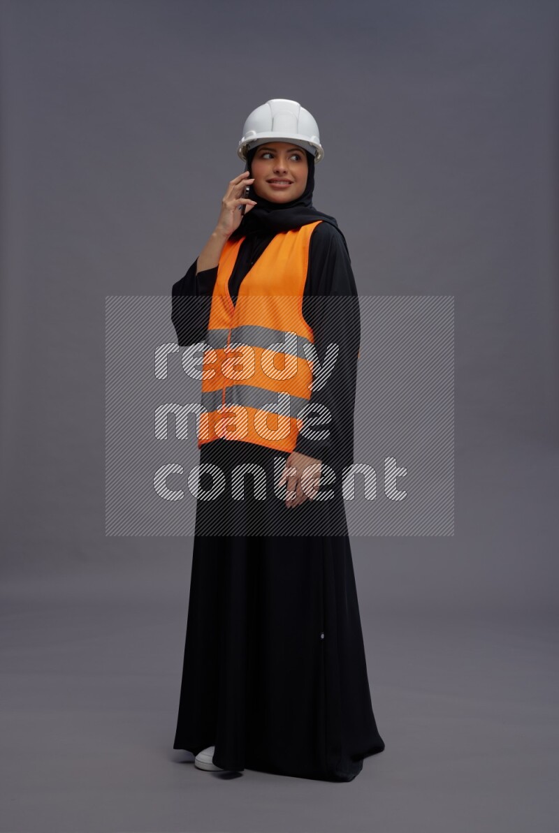 Saudi woman wearing Abaya with engineer vest standing talking on phone on gray background
