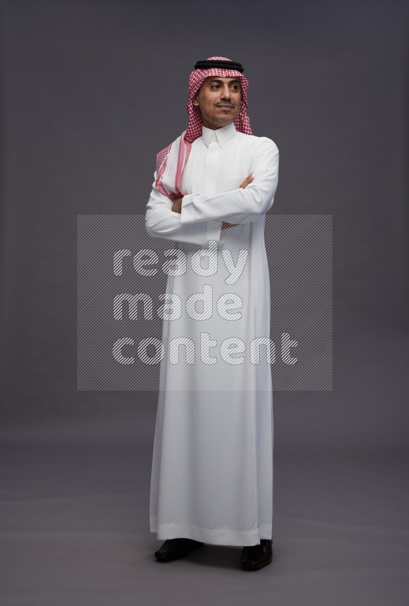 Saudi man wearing thob and shomag standing with crossed arms on gray background